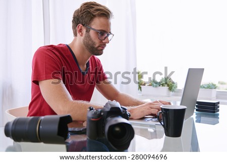 Young professional photographer busy working on his laptop while working from his home studio on his glass desk, while he wears his red shirt and glasses