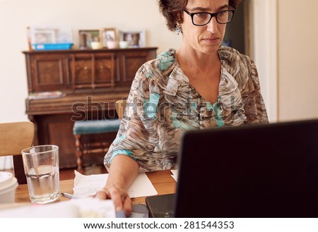 Older entrepreneurial woman busy working in her home office on her new laptop while starring seriously at her screen, with a glass of water to her right