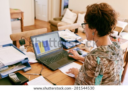 Entrepreneurial business woman busy inputting data from a list onto her new laptop computer from her home office, while sitting at her wooden desk