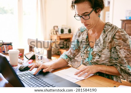 Older business woman busy entering digits on her new laptop computer from a printed list that she\'s reading while wearing glasses