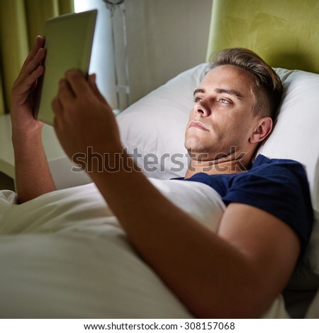 Tired young entrepreneur lying in bed using his tablet to check up his stock trends on last time before sleeping