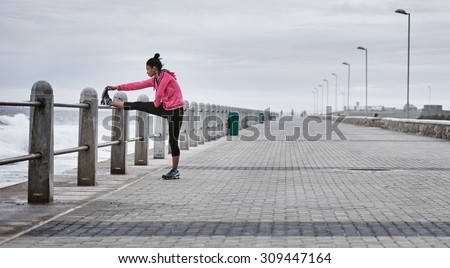 Wide shot landscape image of a mixed race female professional runner wearing a pink sweat shirt busy stretching her quads on a ocean side promenade railing