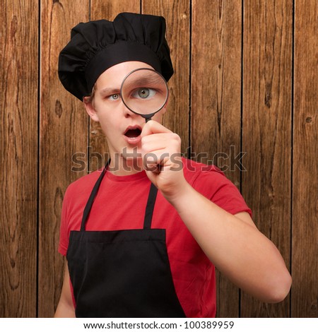 portrait of a young cook man looking through a magnifying glass against a wooden wall