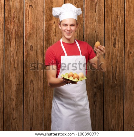 portrait of a young cook man holding box of eggs against a wooden wall