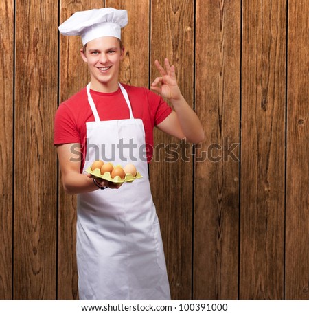 portrait of a young cook man holding a box of eggs and doing a good gesture against a wooden wall