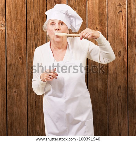 portrait of a cook senior woman with a wooden spoon in her mouth against a wooden wall