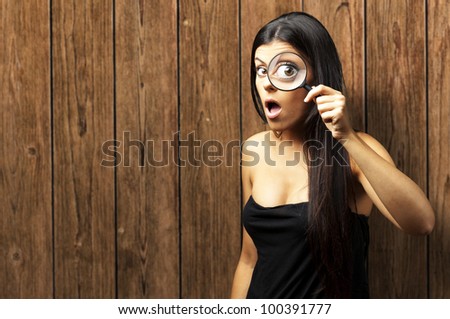 Young woman surprised looking through a magnifying glass against wooden wall