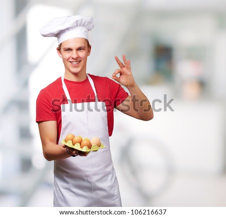 portrait of a young cook man holding an egg box and doing a good gesture indoor