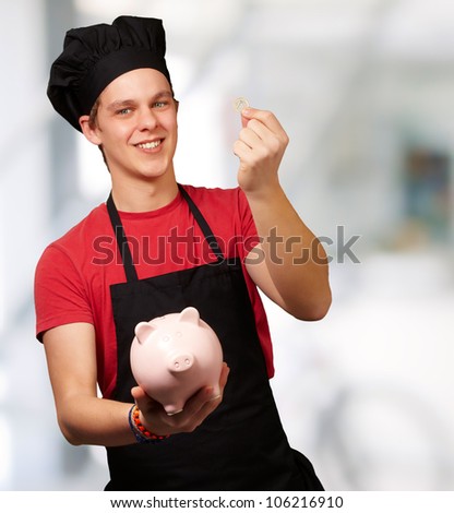 portrait of a young cook man holding a euro coin and a piggy bank indoor