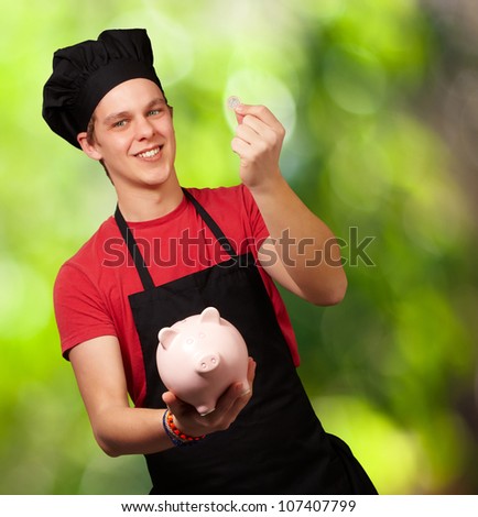 portrait of a young cook man holding a euro coin and a piggy bank against a nature background