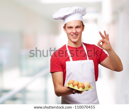 portrait of young cook man holding egg box and doing good gesture against abstract background