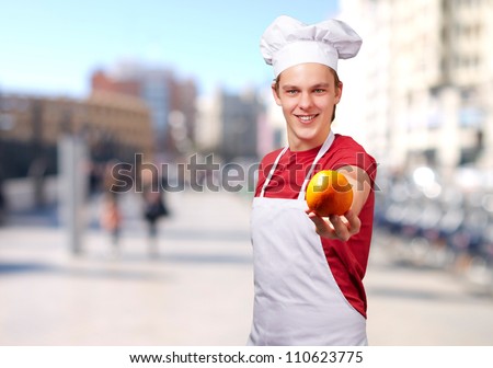 portrait of young cook man offering orange at city