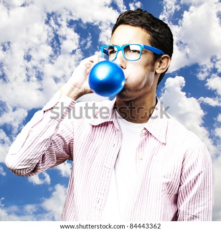 portrait of young man inflating a blue balloon against a sky background