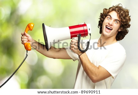 portrait of young man shouting with megaphone and talking on vintage telephone against a nature background