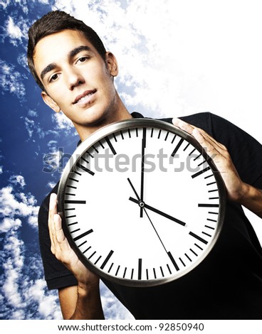 portrait of young man holding a clock with his hands against a cloudy sky background