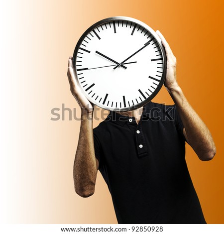 young man covering his face with a clock over orange background