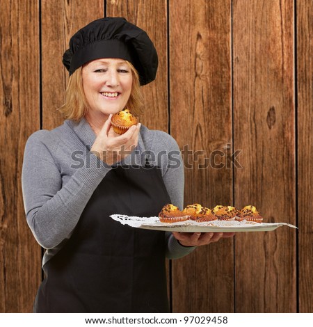 portrait of middle aged cook woman holding a homemade muffin against a wooden wall