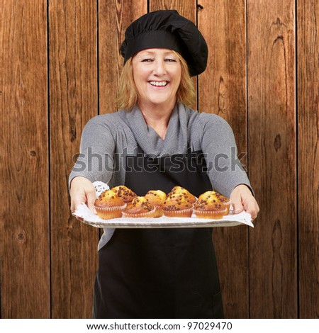 portrait of cook woman showing a homemade muffins tray against a wooden wall
