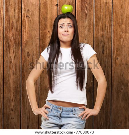 portrait of young woman holding green apple on her head against a wooden wall