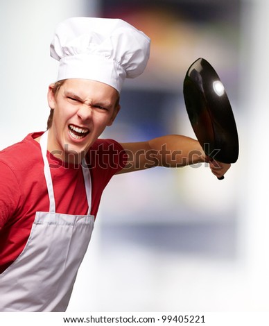 portrait of angry young cook man hitting with pan indoor