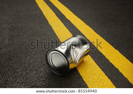 Close up of a dented, shiny, metal can sitting on an black asphalt road with yellow stripes