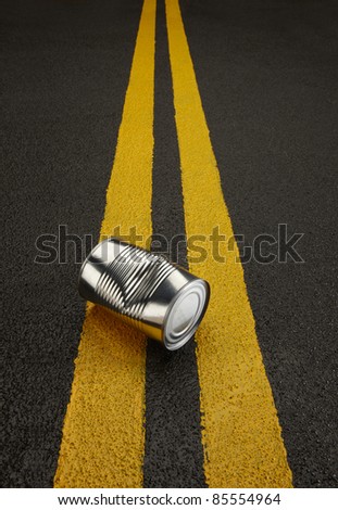Close up of a shiny dentedl can sitting on a black asphalt road with yellow stripes.