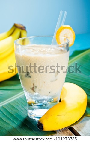 Creamy banana smoothie blended with fresh yoghurt in a glass with ripe bananas and a banana leaf