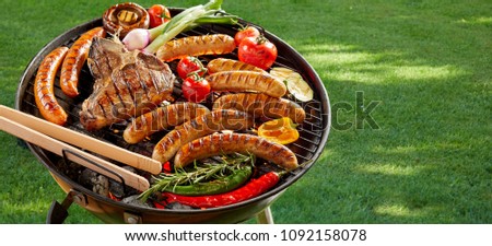 Meat and vegetables grilling on an outdoor portable barbecue on fresh green summer grass with pork sausages, beef steak, bell peppers, chili, onion, zucchini and tomato, banner format with copy space