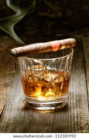 Burning Cuban cigar resting on glass of whisky on ice on an old wooden surface symbolic of masculinity