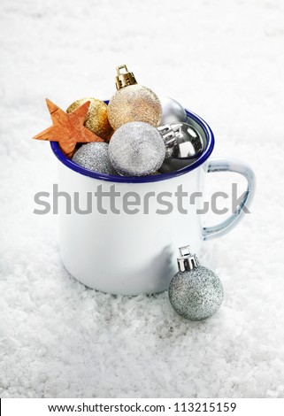 Christmas decorations in an enamelled tin mug on snow which is full to overflowing with baubles and stars