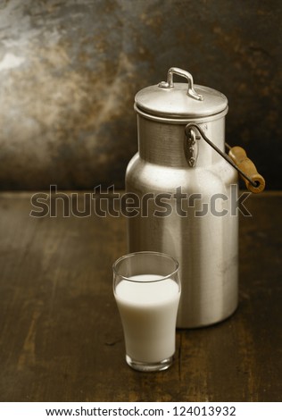 Metal milk can with a glass of fresh milk standing together on an old rustic wooden table in front of a grungy stained wall