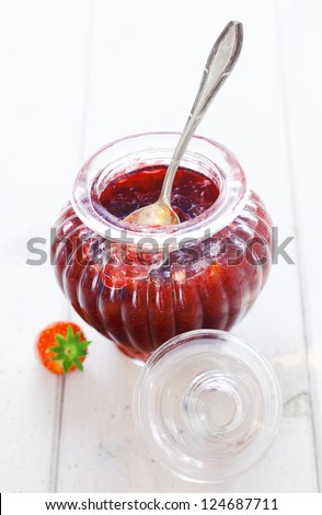 Delicious homemade strawberry preserve in an elegant fluted glass jar with a silver serving spoon on a white background
