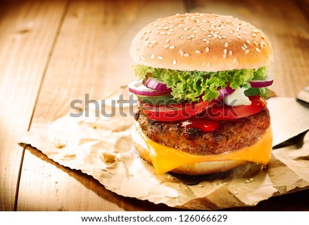 Delicious Cheeseburger Stacked High With A Juicy Beef Patty, Cheese, Fresh Lettuce, Onion And Tomato On A Fresh Bun With Sesame Seed Standing On Brown Paper On A Wooden Tabletop With Copyspace