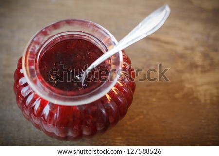 Overhead view of a decorative fluted glass jar filled with fresh fruity strawberry jam and a silver serving spoon on a textured wooden surface with copyspace