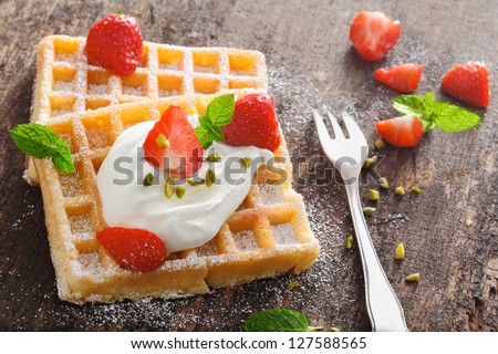 Golden waffle topped with fresh diced strawberries and a dollop of whipped cream sprinkled with sugar and served with a fork on a rough textured old wood surface