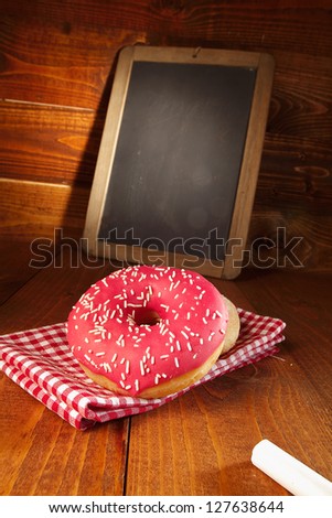 Freshly baked donuts glazed with red icing and sprinkles served on a red and white checked cloth in front of a small blank chalkboard