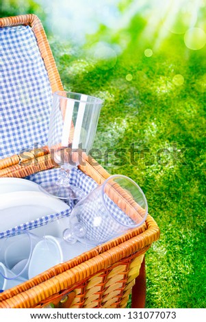 Fresh bright picnic basket with a blue and white checked country lining open to display glasses and cups against lush greenery