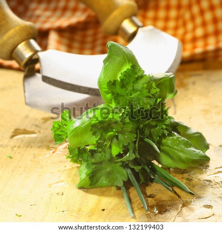 Bouquet garni of fresh herbs including basil, crinkly leaf parsley and chives lying on a wooden table in front of a chopping blade