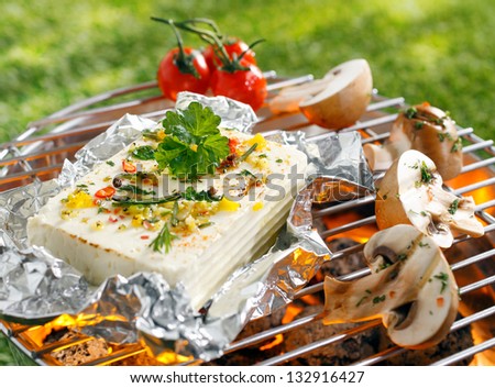 Large portion of seasoned halloumi or feta cheese grilling in tin foil over hot coals in a barbecue outdoors on green grass