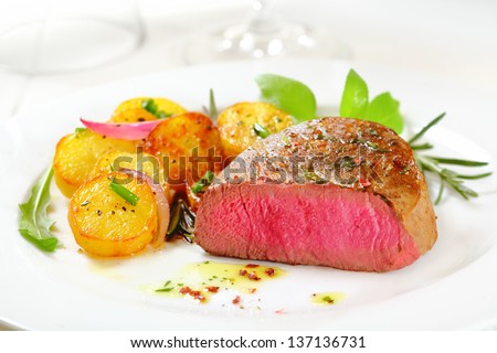 Cut through portion of delicious bloody rare to medium rare fillet steak served with crispy roasted potatoes, fresh rosemary and rocket on a white plate