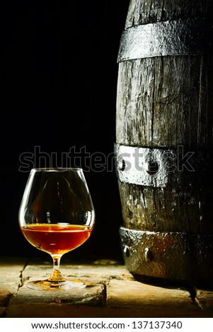 Cognac snifter with glowing golden cognac standing on old flagstones or tiles alongside an old oak barrel in a dark cellar, conceptual of aged matured cognac with copyspace