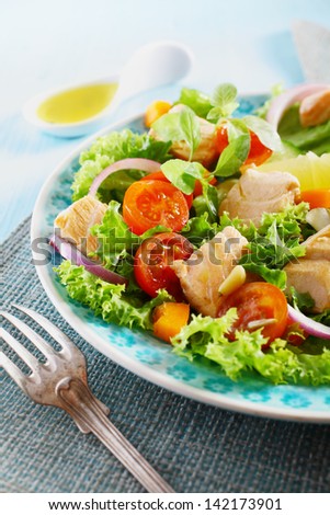 Healthy Mediterranean salad with chicken, pine-seeds, lettuce, cherry-tomatoes, onions, basil and lemon on a blue plate, with a fork in the foreground and an olive oil spoon in the background