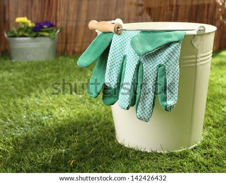 Green textile gardening gloves hanging over the rim of a metal bucket standing on a green lawn in summer sunshine, conceptual image