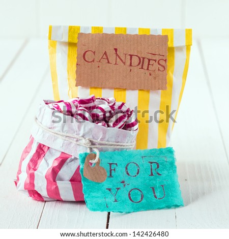 Close-up of two colorful bags with white, yellow and pink stripes filled with candies with romantic message in tags meaning, Candies For You, over a white wooden background