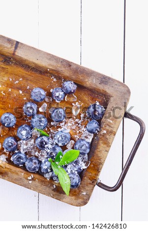 Close-up of ripe blueberries or bilberries and ice in an old grungy wood tray with a small flowery branch over a white wooden table