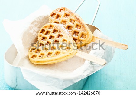 Two heart shaped Belgian waffles on a stick inside a tin can a a paper wrap, sprinkled with cinnamon and sugar