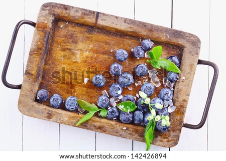 Ripe blueberries or bilberries in an old grungy wood tray with a small flowery branch over a white wooden table