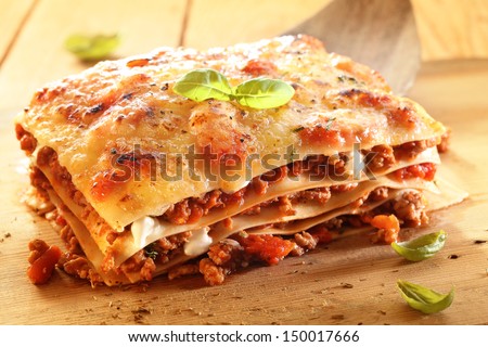 Golden lasagne with meat, tomatoes, cheese sauce and pasta in alternating layers on a wooden board garnished with basil