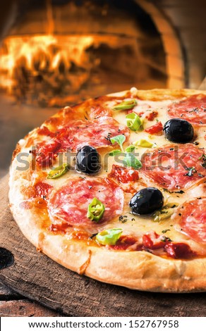 Delicious fresh oven baked spicy salami or pepperoni pizza with melted cheese and olives on a wooden board in front of a roaring fire in a pizza oven at a pizzeria