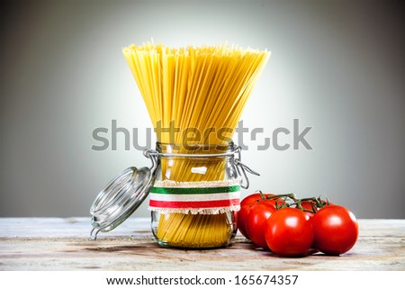 Uncooked dried Italian spaghetti tied with a ribbon in the colours of the national flag - red, white and green - standing in a glass jar with fresh ripe red tomatoes alongside